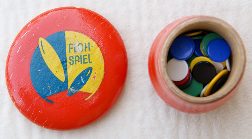 Tucker Tw ID • UNK-164c1 — AGPI ID • G-38349c1 — publisher • (unknown, German) — title • FLOH-SPIEL — notes • Red wooden mushroom container. Yellow and blue illustration on top.